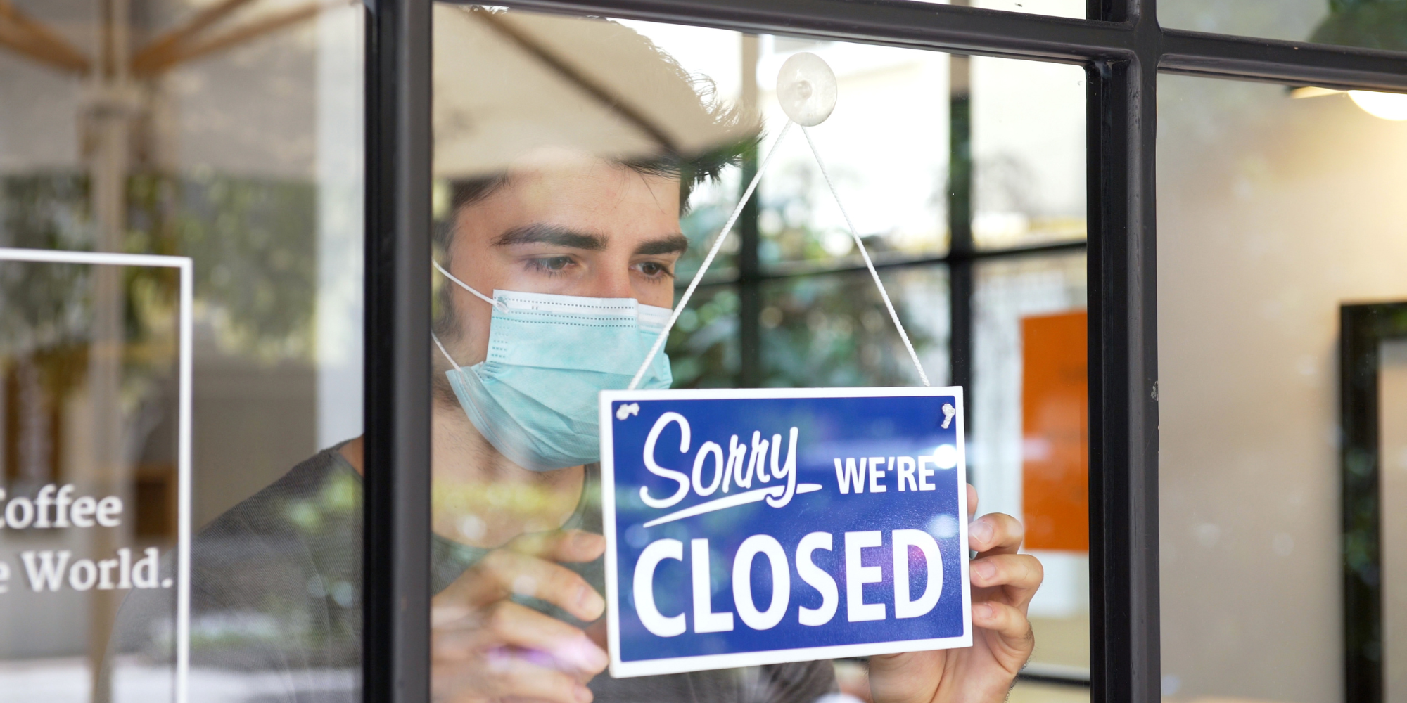 Young masculine, white presenting person with dark hair wearing a blue mask putting up a "Sorry we're closed" sign in a coffee shop window - white text on blue background.