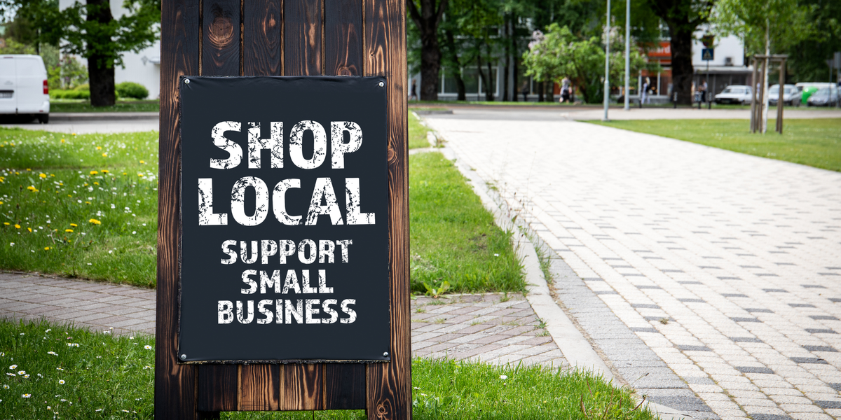 Dark wood board sign on grassy area near grey interlock paths that says in white, all caps, "Shop Local: Support Small Businesses."."