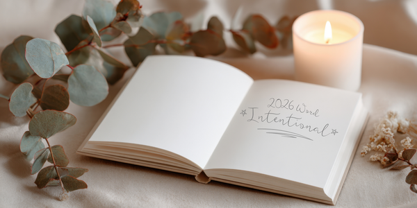Beige journal open to blank pages reading ‘2026 Word Intentional,’ styled with eucalyptus, a candle, and a small white flower.